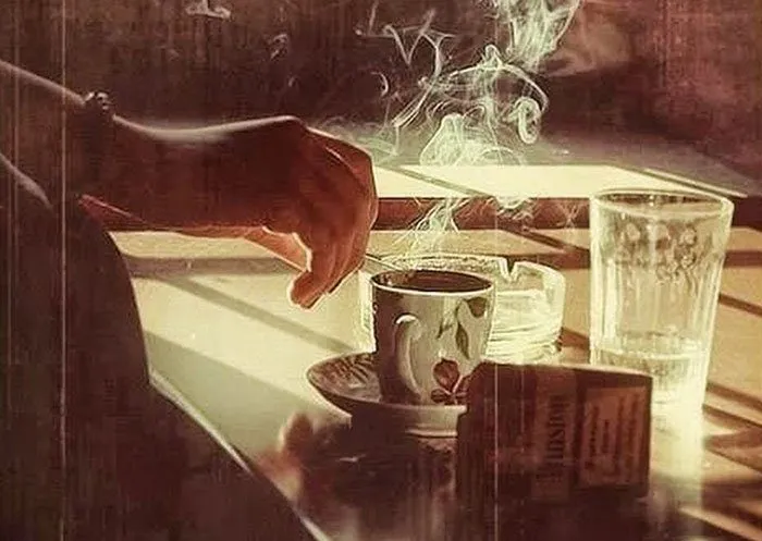 Ly cà phê và điếu thuốc đang cháy dở trên bàn, gợi lên không gian yên tĩnh và trầm tư.